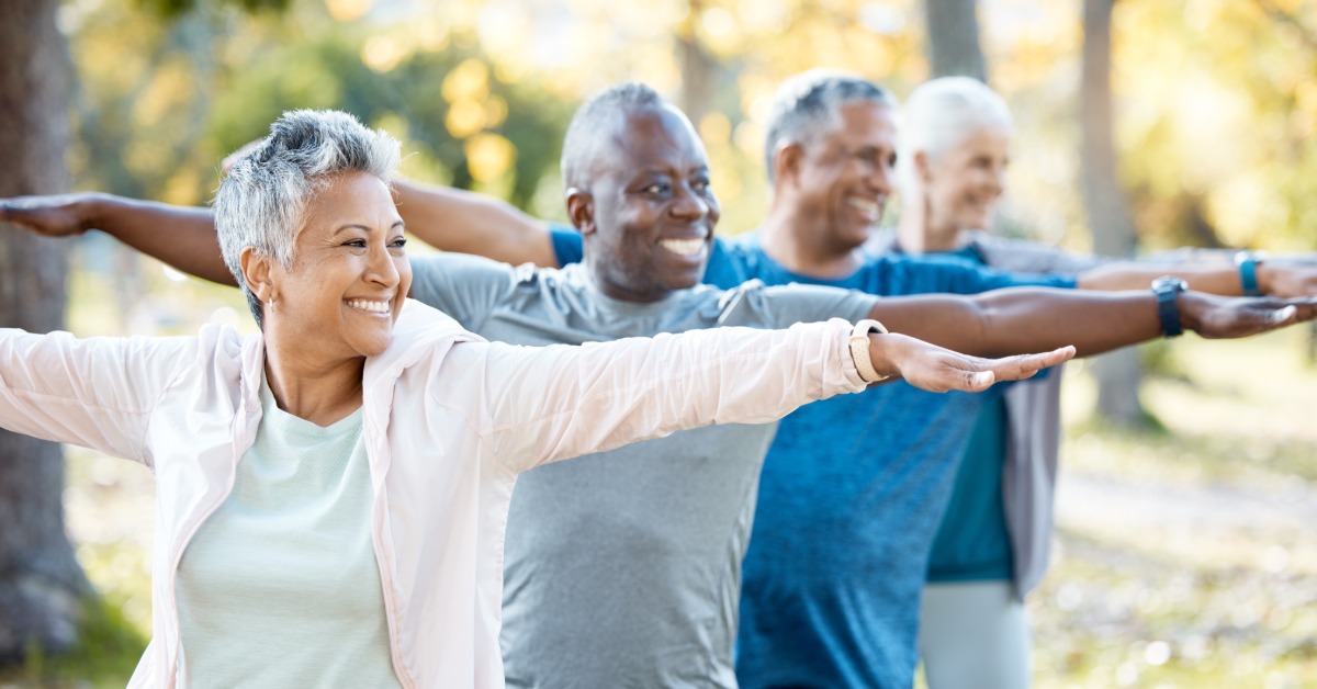 A group of seniors is stretching their arms outdoors in a park while smiling and participating in light exercise.