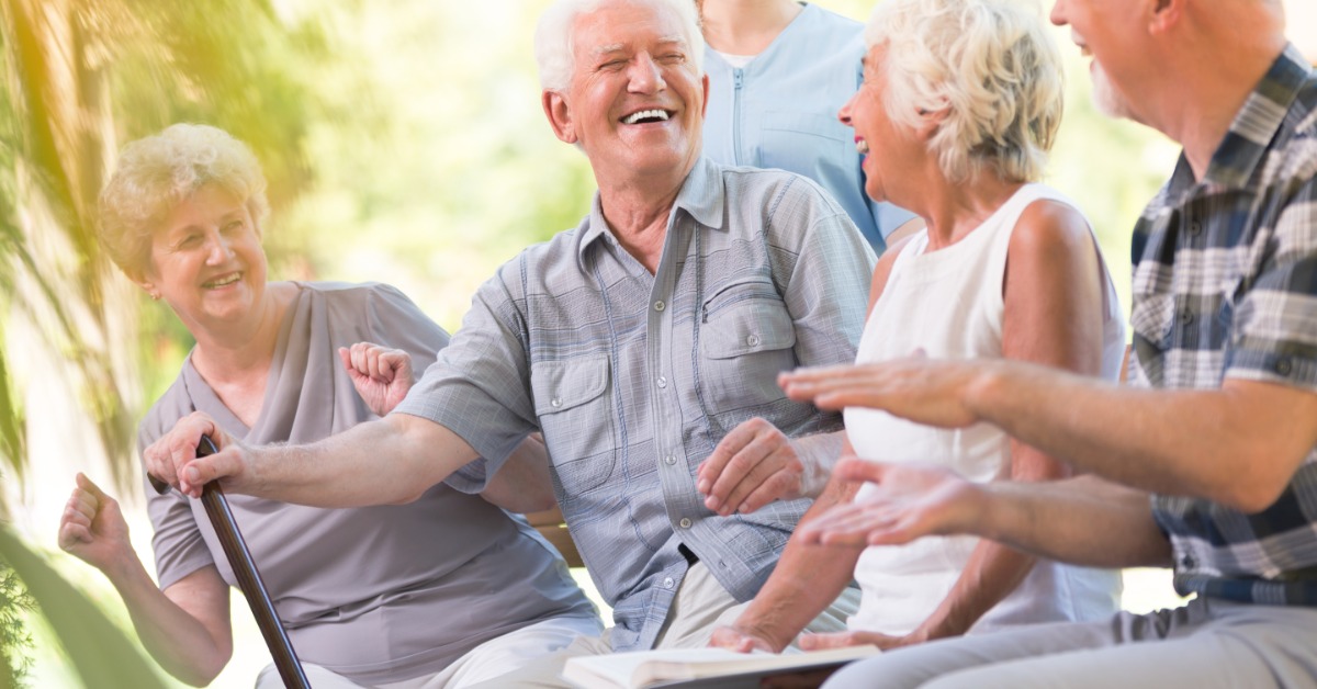 A group of senior adults laughing together on a park bench while chatting and enjoying time outdoors in warm daylight.