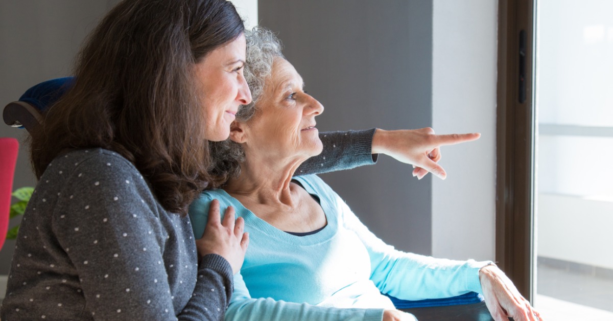 A middle-aged woman sits beside an older woman in a chair and points toward something outside a nearby window.