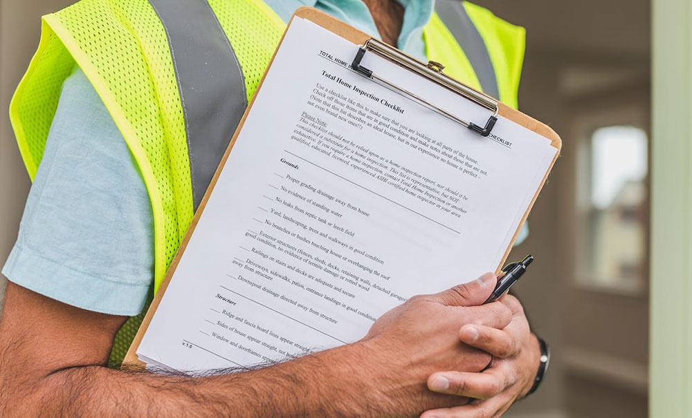 A man in a yellow reflective safety vest holding a pen and a checklist for plumbing and electrical checks in home inspections