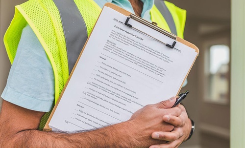 A man in a yellow reflective safety vest holding a pen and a checklist for plumbing and electrical checks in home inspections