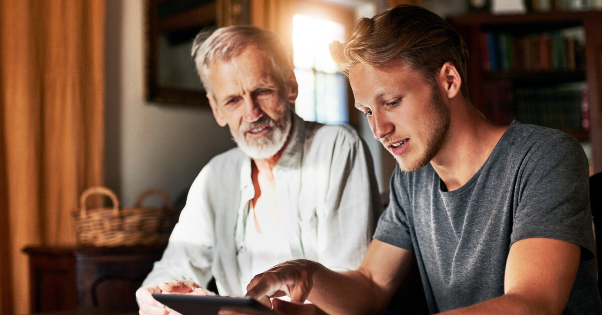 An older man and a younger man looking at a tablet and sitting together at a table with papers and a calculator on it.