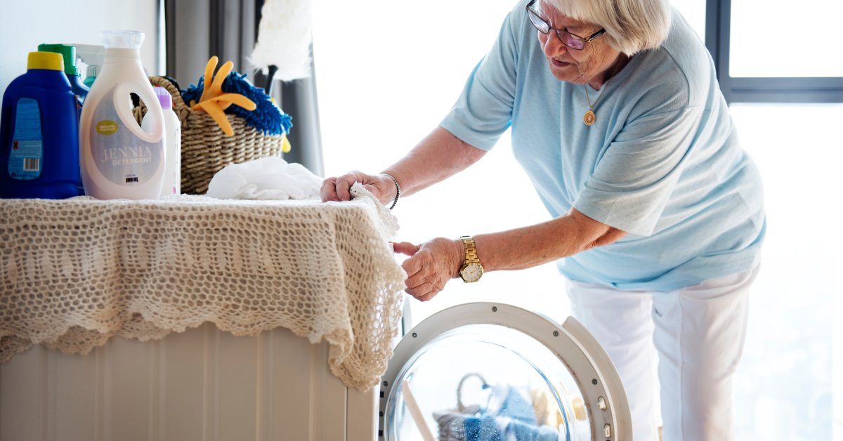 A woman with glasses bends toward an open washing machine with a wicker basket of clothes on the floor beside her.
