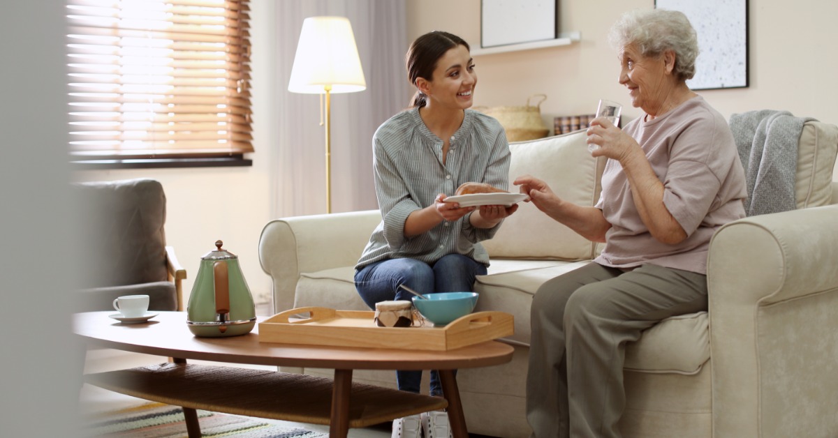 A caregiver consults with an elderly couple on a sofa while taking notes, with a walker nearby in a home setting.