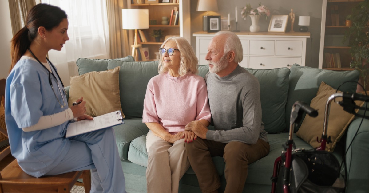 A caregiver serves food to an elderly woman on a sofa while they share a warm, supportive moment at home.