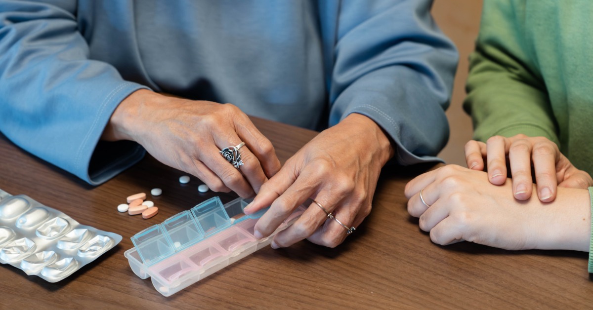 A woman sorts several pills into a medication organizer while another person sits beside her at the table.