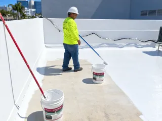 Worker applying white roof coating beside a parapet wall